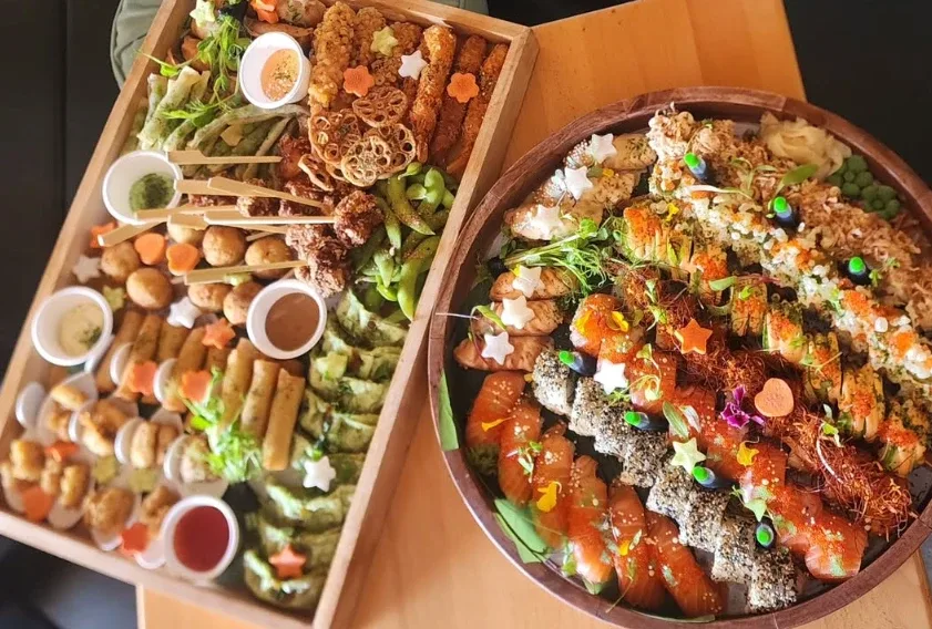 A Kaze Bento catering board of gyoza, spring rolls, crispy chicken, edamame and lotus chips beside a platter of sushi rolls